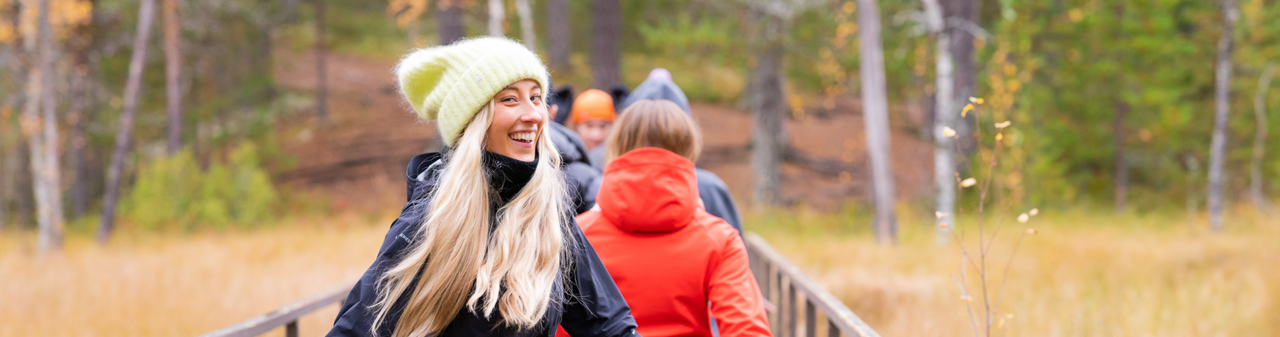 Keltapipoinen nainen katsoo taaksepäin ja hymyilee. Hän on kävelemässä ihmisryhmän kanssa syksyisellä suolla. Hän on jonon viimeisenä.