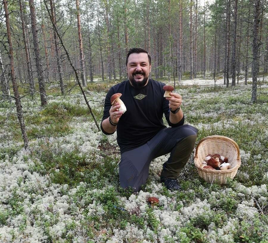 Henkilö sienestää metsässä ja pitää kädessä kahta sientä. Vieressä on sienikori.