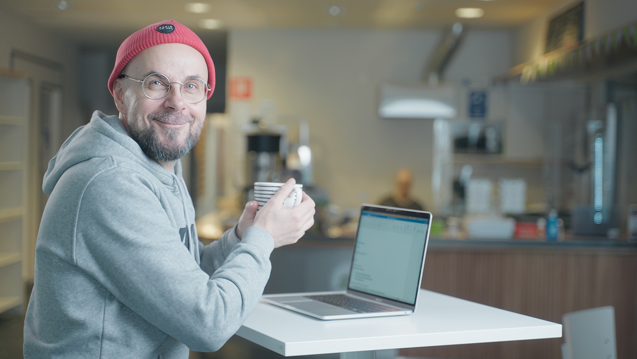 Pipopäinen mies istuu kahviossa kannettavan tietokoneen äärellä kahvikuppi kädessä