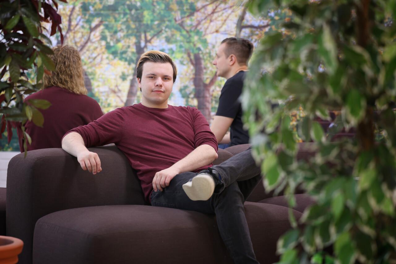 A young man is sitting on a brown sofa. He is wearing a burgundy shirt. Behind him, two people can be dimly seen sitting at the table with their backs to the picture.