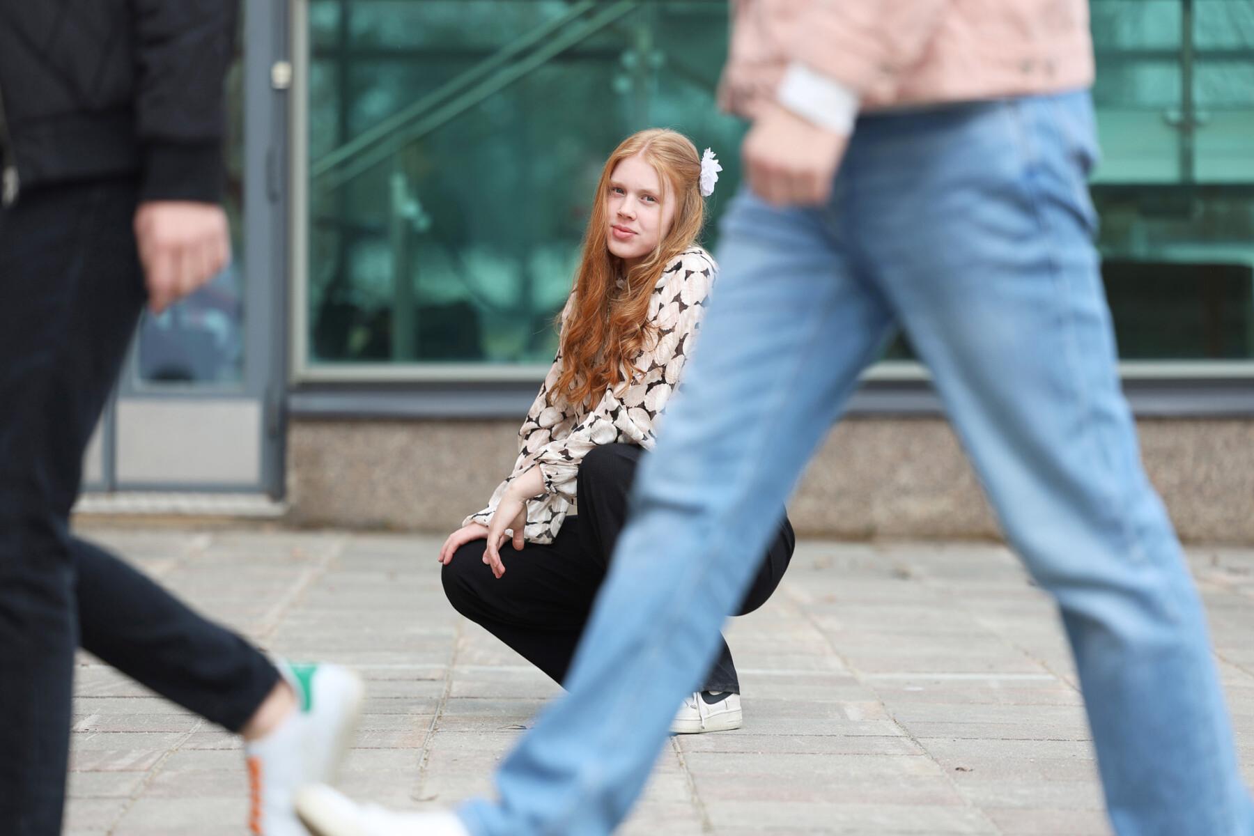 A red-haired girl is squatting on the street. People are walking in front of him. A glass window and a door can be seen in the background.