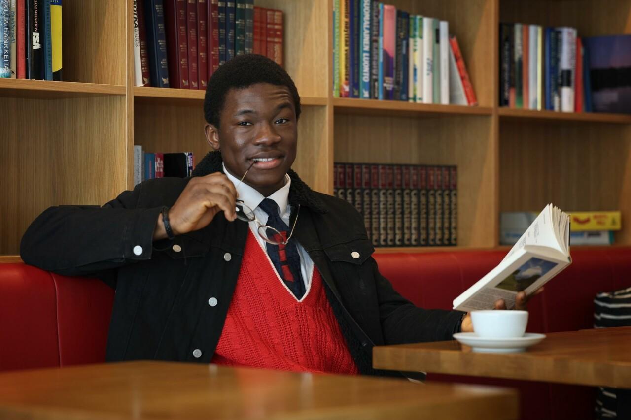 a man reading a book at a cafe table. he has glasses in his hand and a cup of coffee on the table. A bookshelf can be seen in the background.