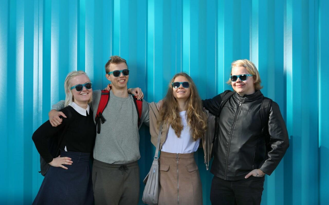 2 girls and 2 boys are standing front of petrol colored metalic wall. They ar wearing sun classes and smiling.