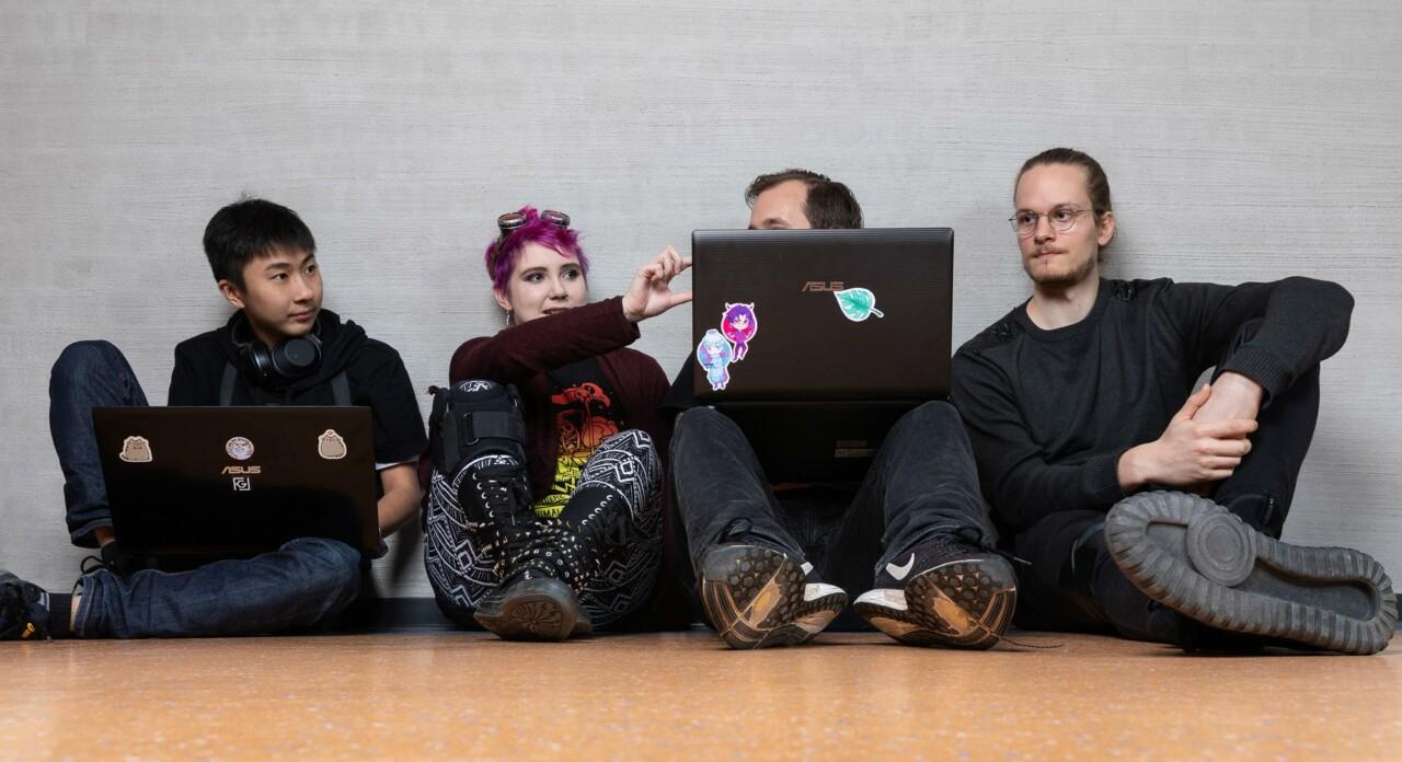 four young people sitting leaning against the wall, two of them have a computer on their lap