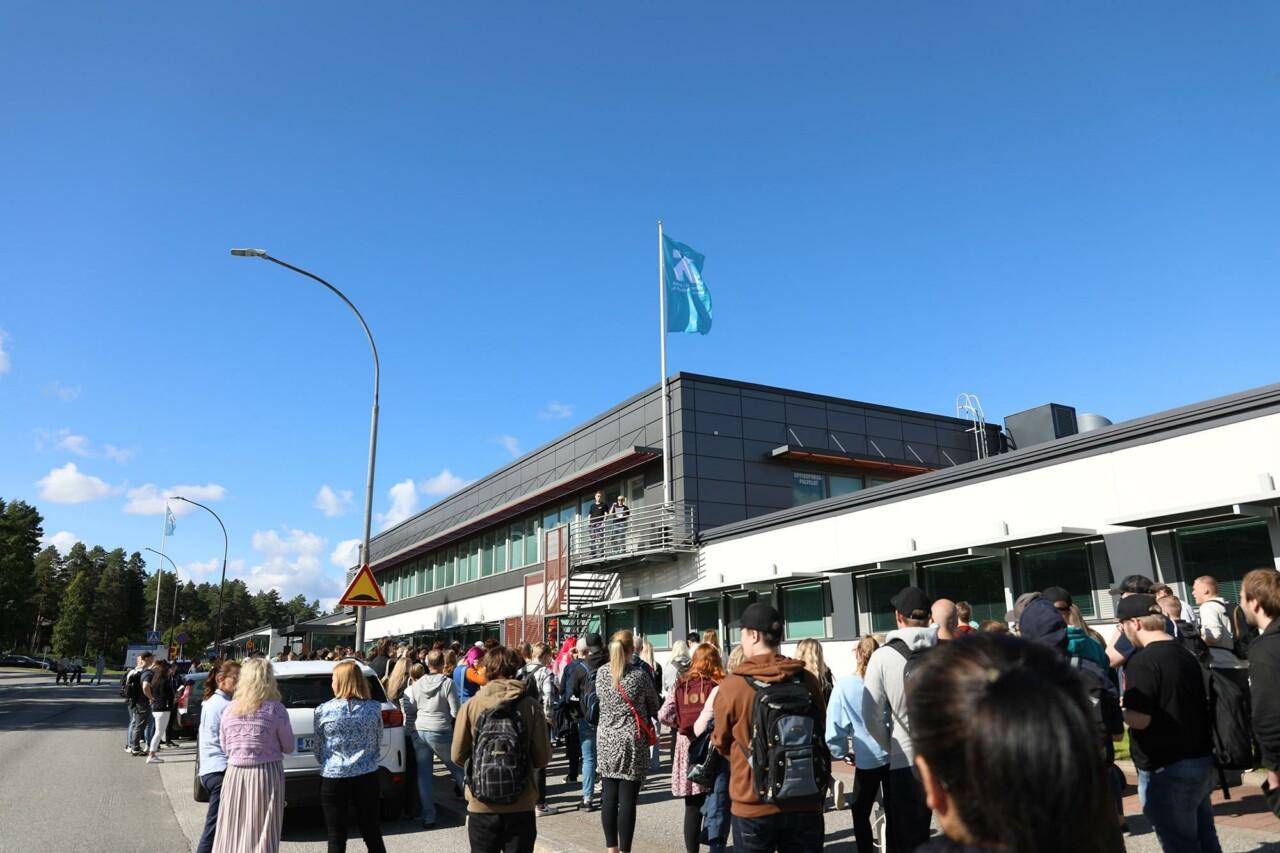 Joukko opiskelijoita on kokoontunut talon eteen. He katsovat ylöspäin parvekkeelle, jossa seisoo useampi henkilö. Parvekkeen vieressä on lippitanko jos liehuu lippu.