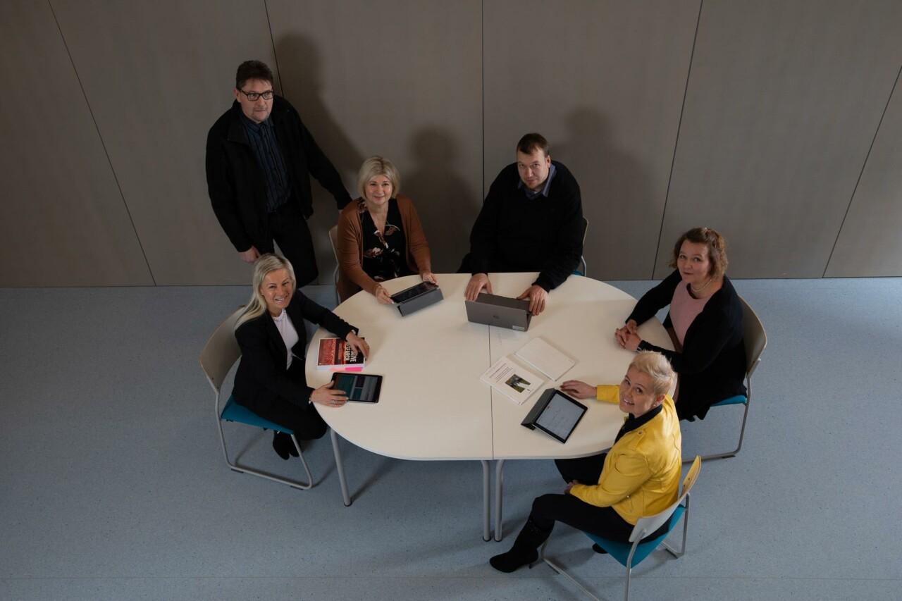 Five people sit around a table and they look up. Behind them stands a man with glasses.