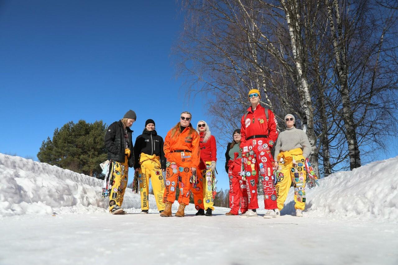 Seven young students are standing on a winter road, they are wearing student overalls. The overalls are yellow, red and orange