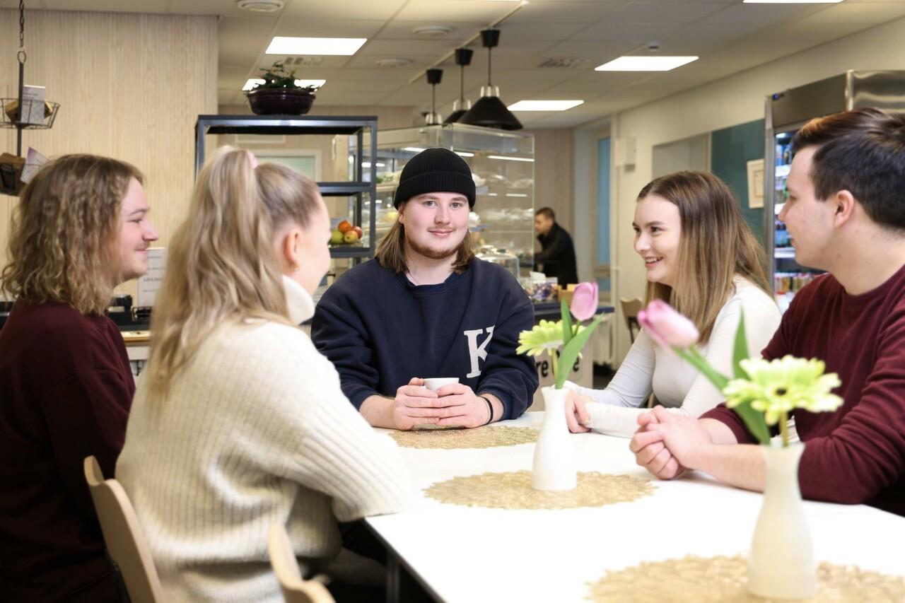 Viisi nuorta opiskelijaa istuu kahvilan pöydän ääressä ja keskustelee.