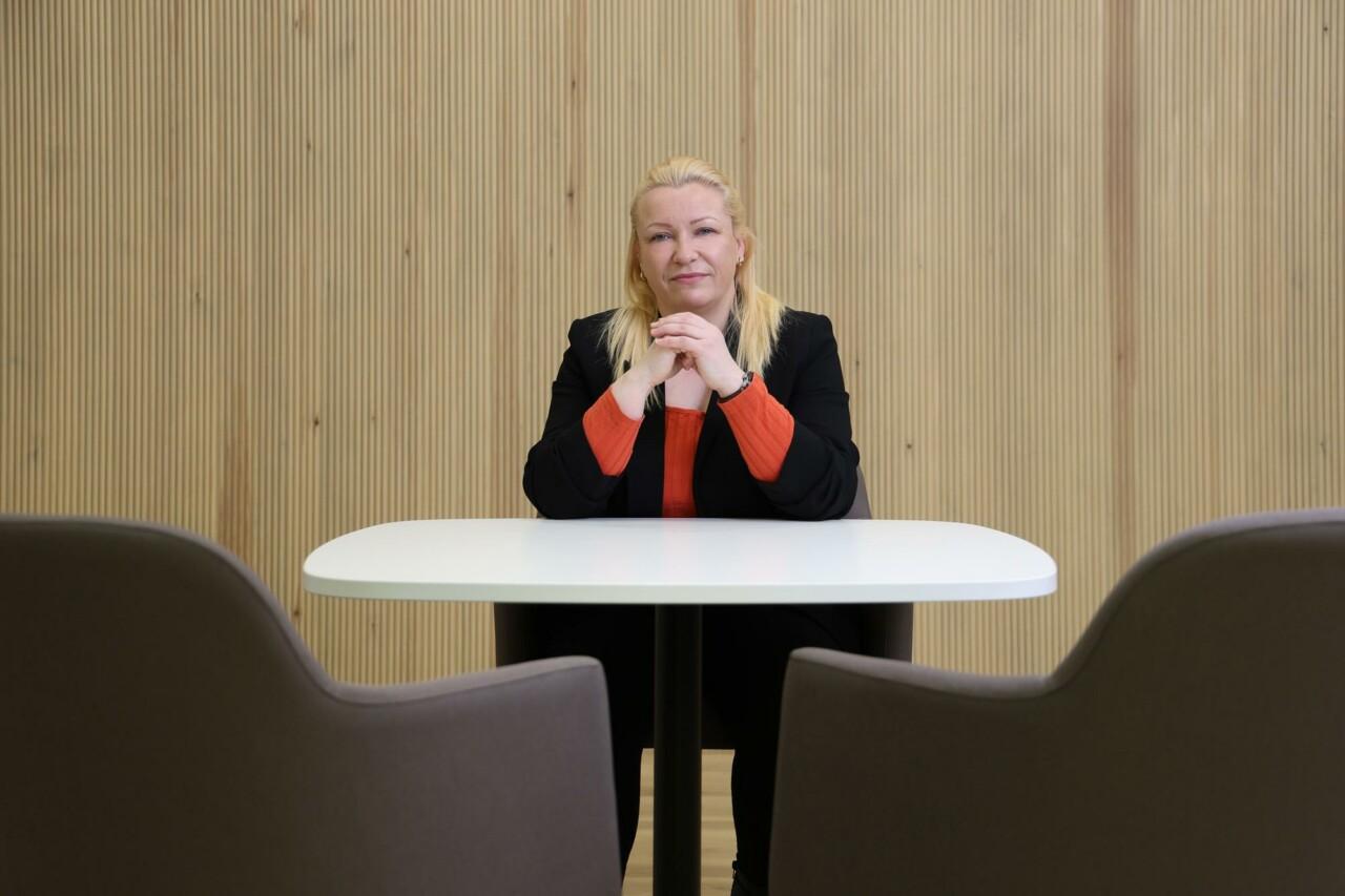 A blond haired woman sitting at a table. She is wearing orange shirt and a black blazer.