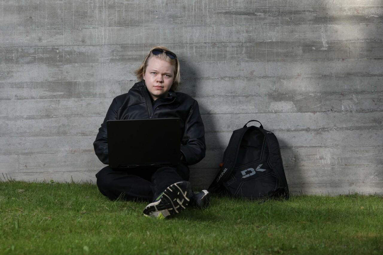 A young man sits on the grass next to the concrete wall with a laptop in his lap and looks ahead