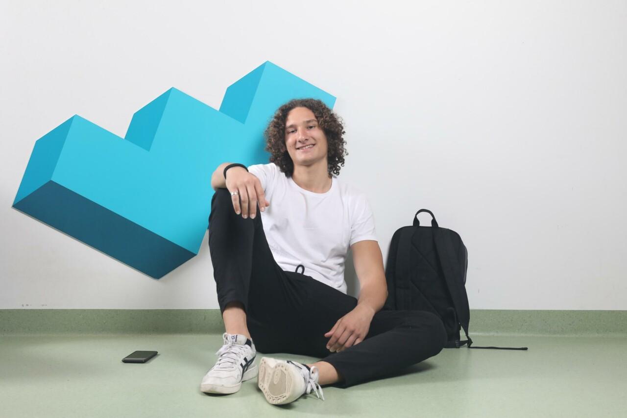 A boy with curly hair is sitting on the floor in front of a white wall, he has a backpack next to him.