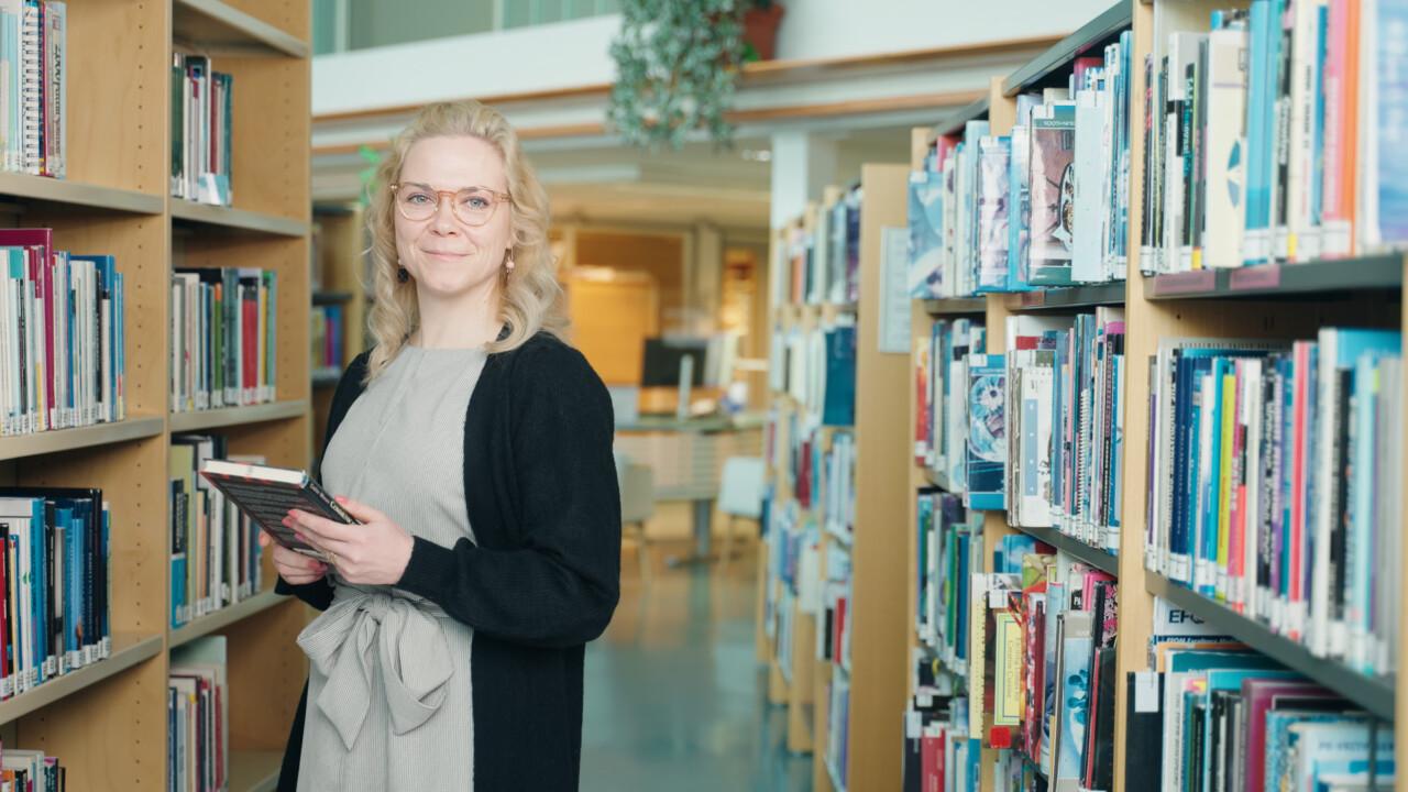 Vaaleahiuksinen nainen seisoo kirjastohyllyjen välissä ja pitää kädessään kirjaa. Naisella on silmälasit, vaaleanruskea mekko ja musta neuletakki.