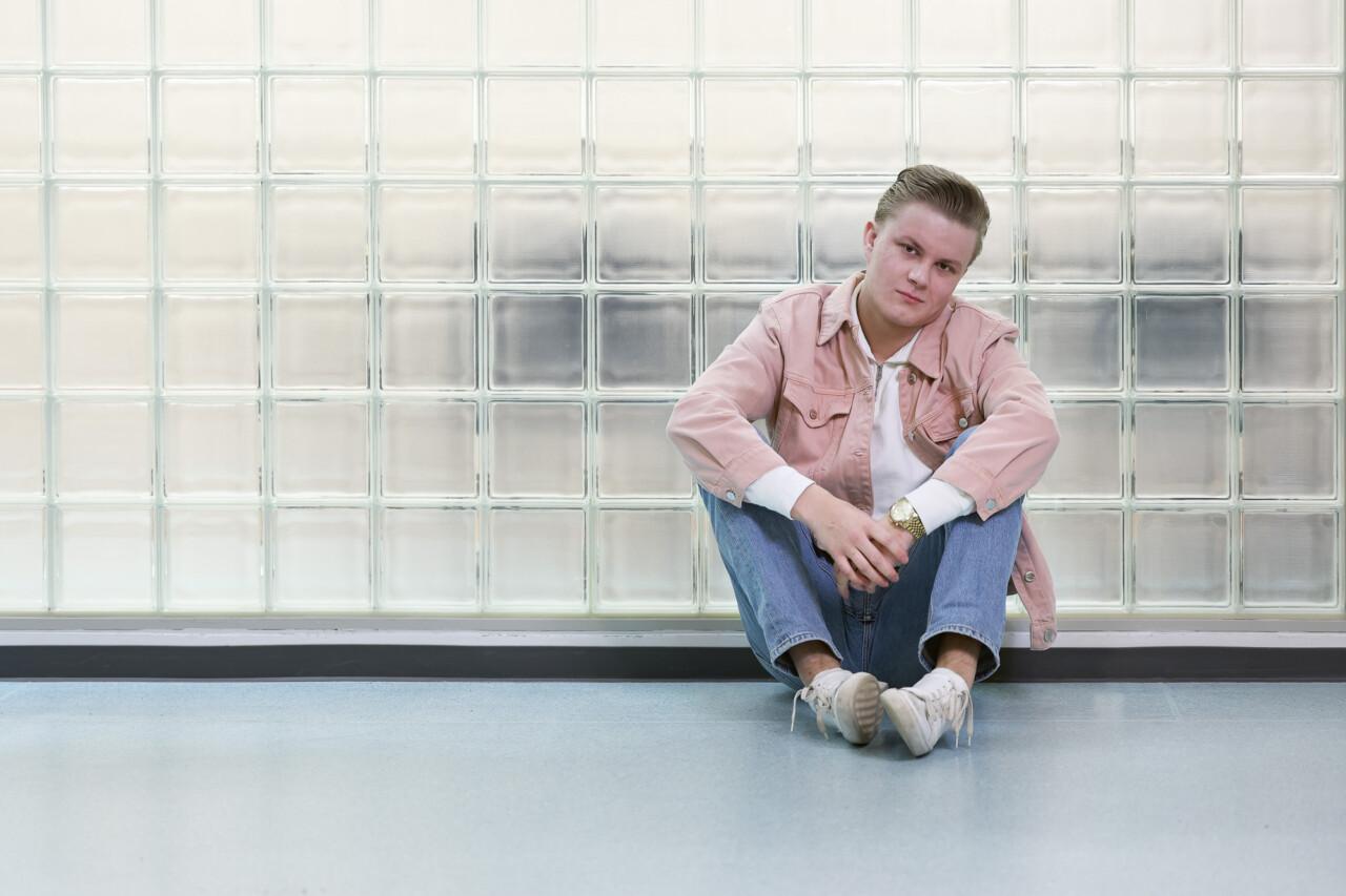 Nuori mies vaaleanpunaisessa paitapuserossa istuu lattialla lasitiiliseinän edessä.