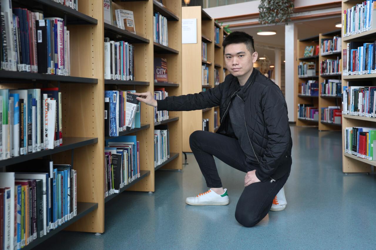 Aasialaistaustainen mies on polviasennossa kirjaston hyllyn edessä ja ottaa kirjaa hyllystä.