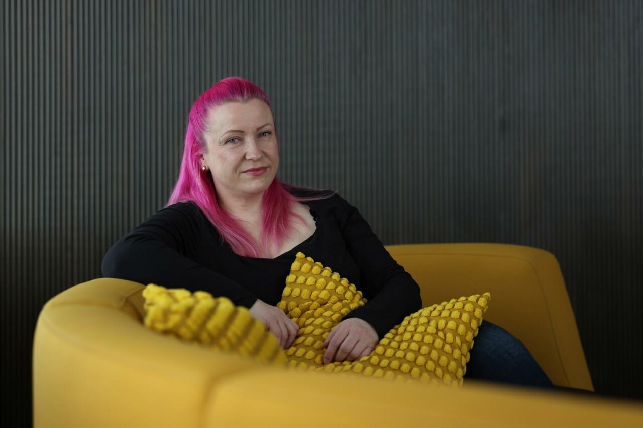 Pinkkihiuksinen nainen istuu keltaisella sohvalla tyyny sylissään.