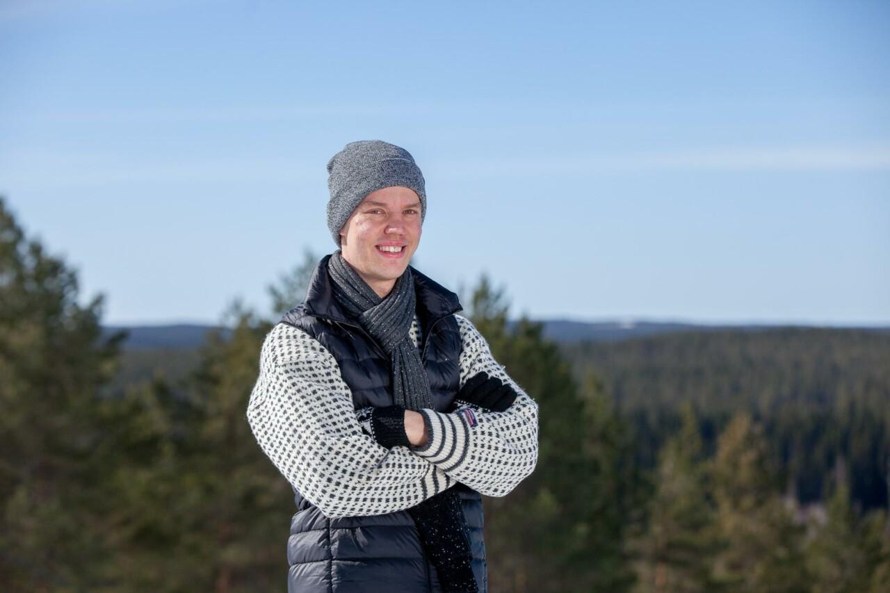 A young man stands outside and smiles. Behind you can see a forest and a hill landscape