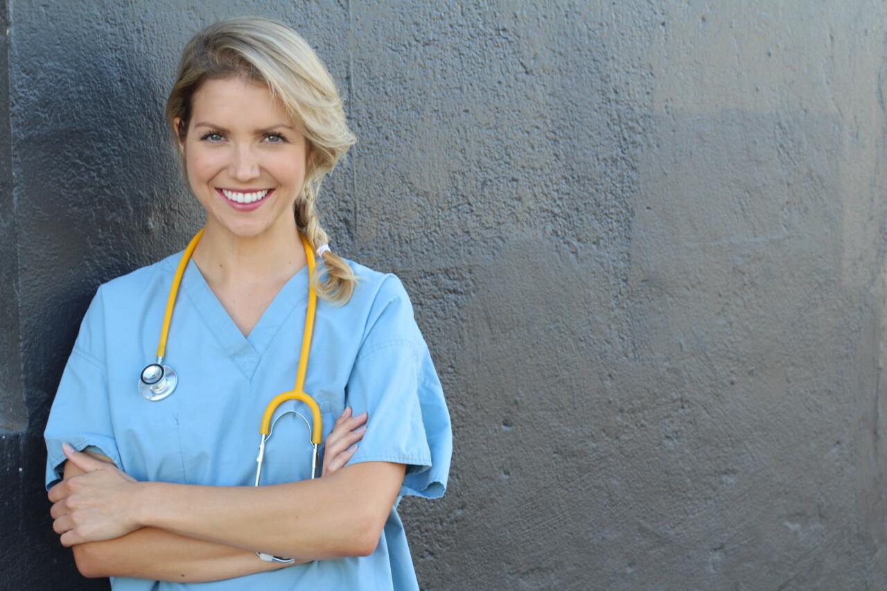 A female nurse leans against the wall with a stethoscope around her neck