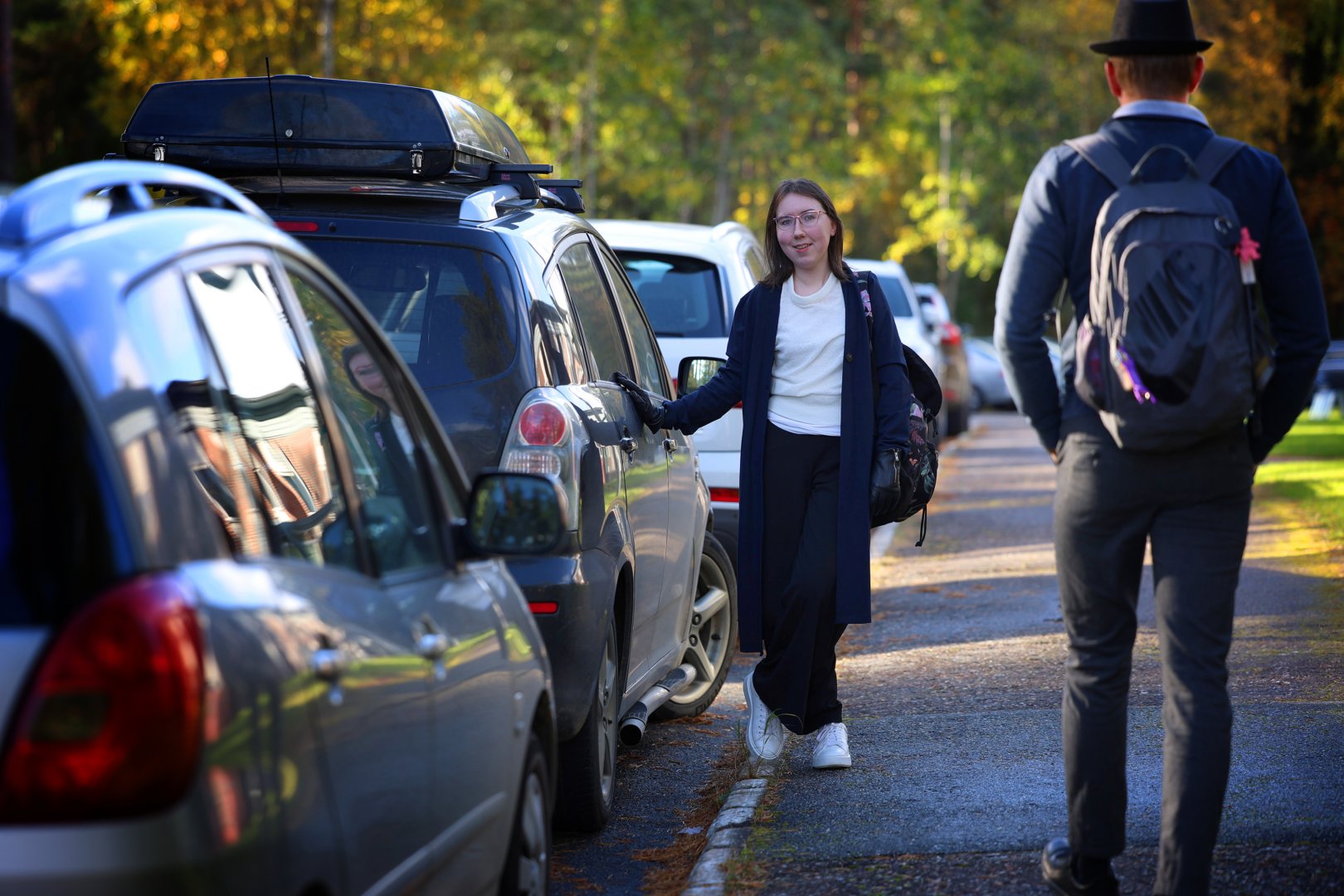 Nainen nojaa kädellään harmaaseen autoon katsoen kameraan. Hänen vierestään kävelee leirihattuinen mies selkä kameraan päin.