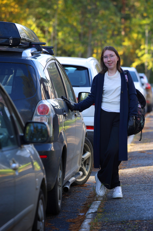 Nainen nojaa kädellään harmaaseen autoon katsoen kameraan. Hänen vierestään kävelee leirihattuinen mies selkä kameraan päin.