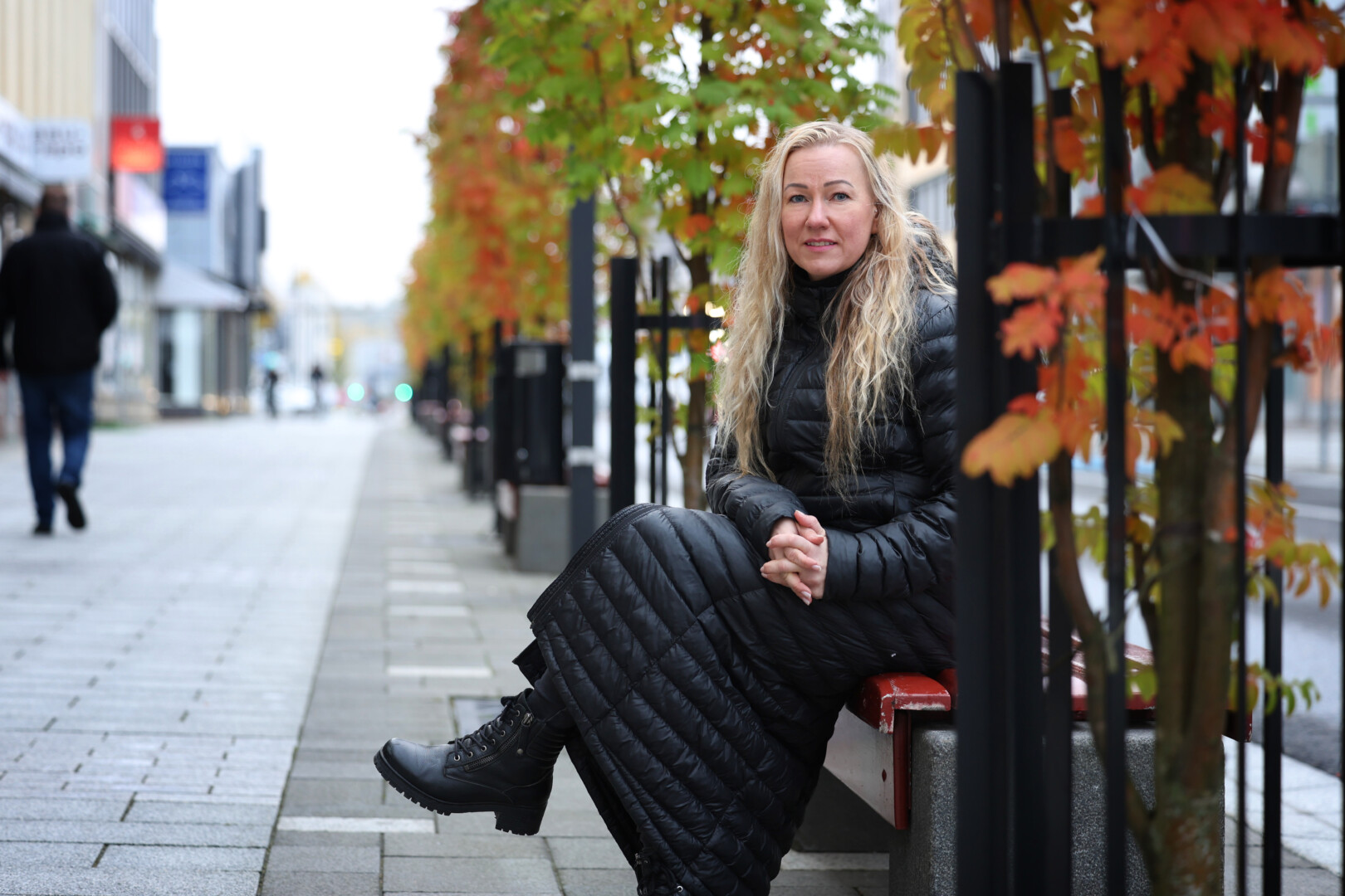 A blonde woman sitting on a bench outside with a long coat