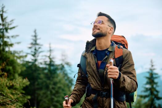 A happy hiker with a hiking pole in his hand and a backpack on his back. Forest in the background. Picture: Drazen / Adobe Stock