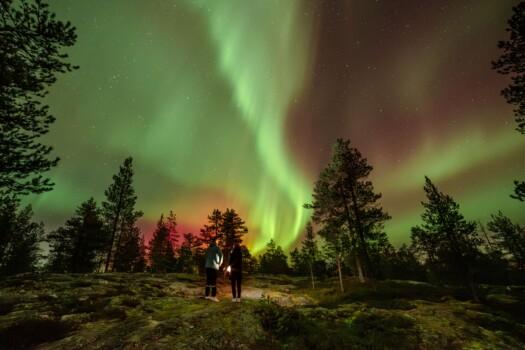 Two people standing in the forest in the middle of night with green northern lights in the sky. Photo: Roni Lukkarinen.