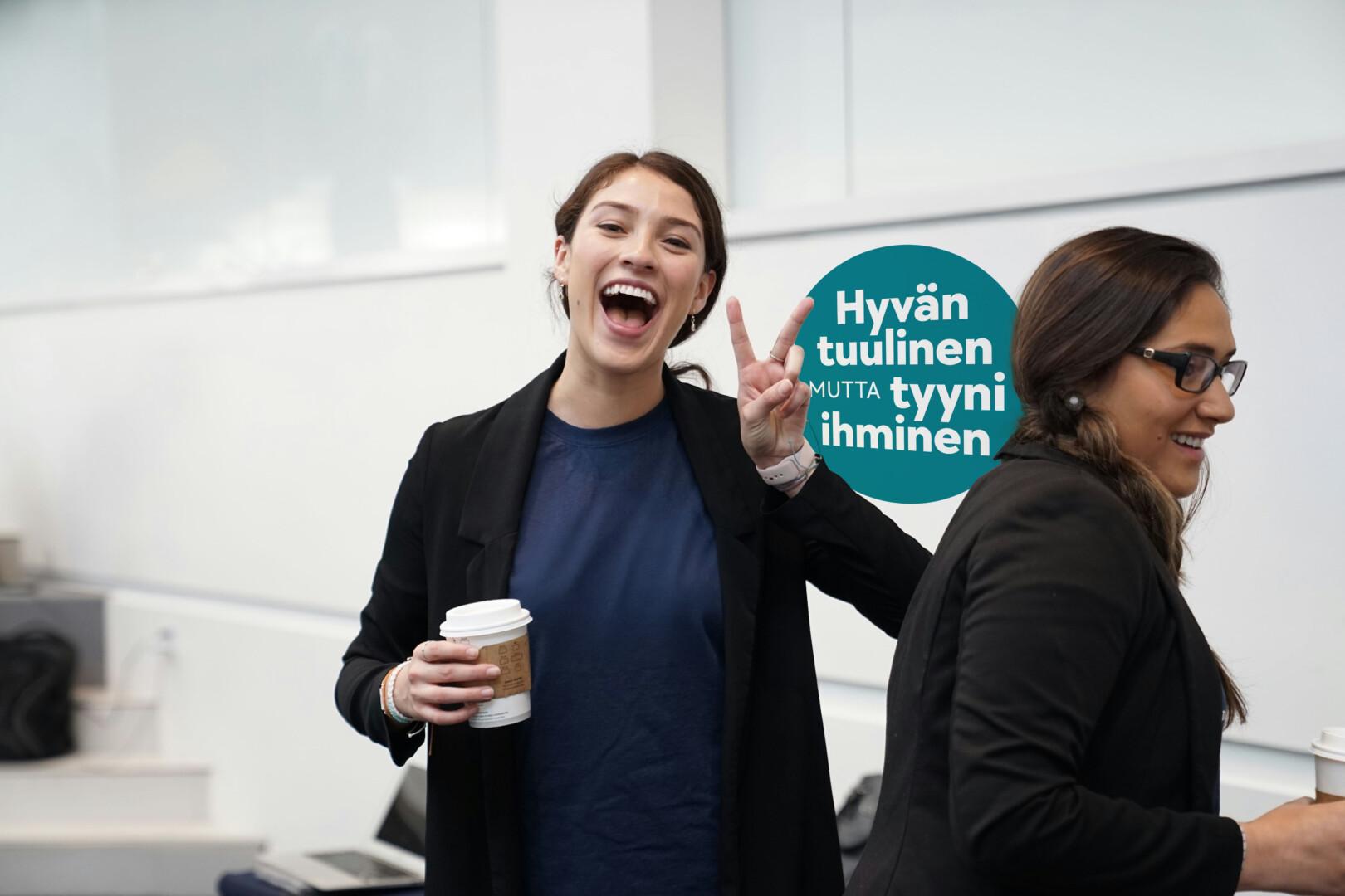 Kaksi naishenkilöä, toinen hymyilee kameraan päin. Naisten välissä on värillinen pallo, jossa lukee "Hyvän tuulinen, mutta tyyni ihminen"