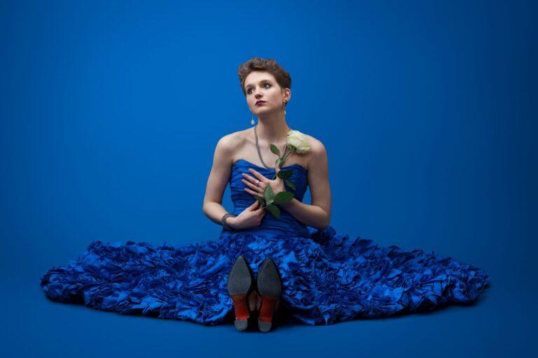 Singer Sara Emilia. She is sitting on the floor and wearing a blue dress. She holds a rose in her hands.