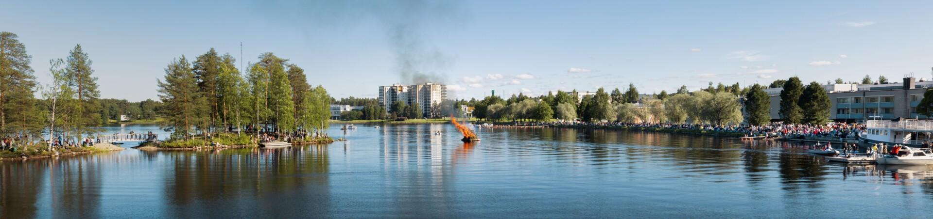 Copilot said: Photograph of Kajaani during Midsummer. On the left, a bridge and an island can be seen; in the middle of the river, a Midsummer bonfire is burning; and on the right, a harbour area filled with people celebrating. Photo: Ann Bernevega.